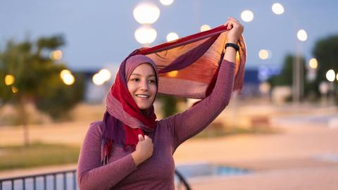 Eine junge Frau lächelt auf der Straße während sie eine Stoffstreifen ihres Kopftuchs in das Laternenlicht hält.
