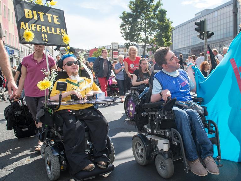 Drei Demonstranten bei der "Pride Parade 2014" tragen Transparente und Schilder. Auf einem Schild steht: "Bitte auffallen!". Zwei Demonstranten fahren in Rollstühlen, einer läuft. Im Hintergrund weitere Demonstranten.
