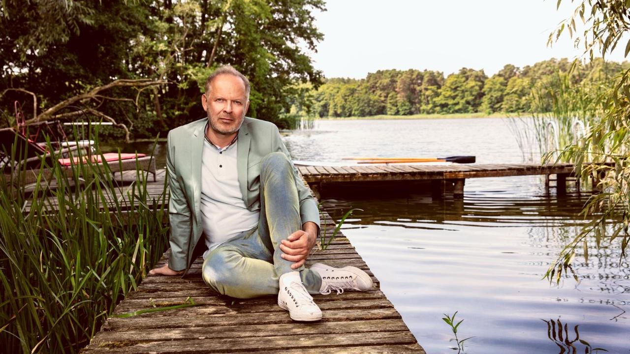 Axel Milberg sitzt bekleidet mit Jeans, Sakko und Turschuhen auf einem Bootssteg. Im Hintergrund ein See und Ruderboote.