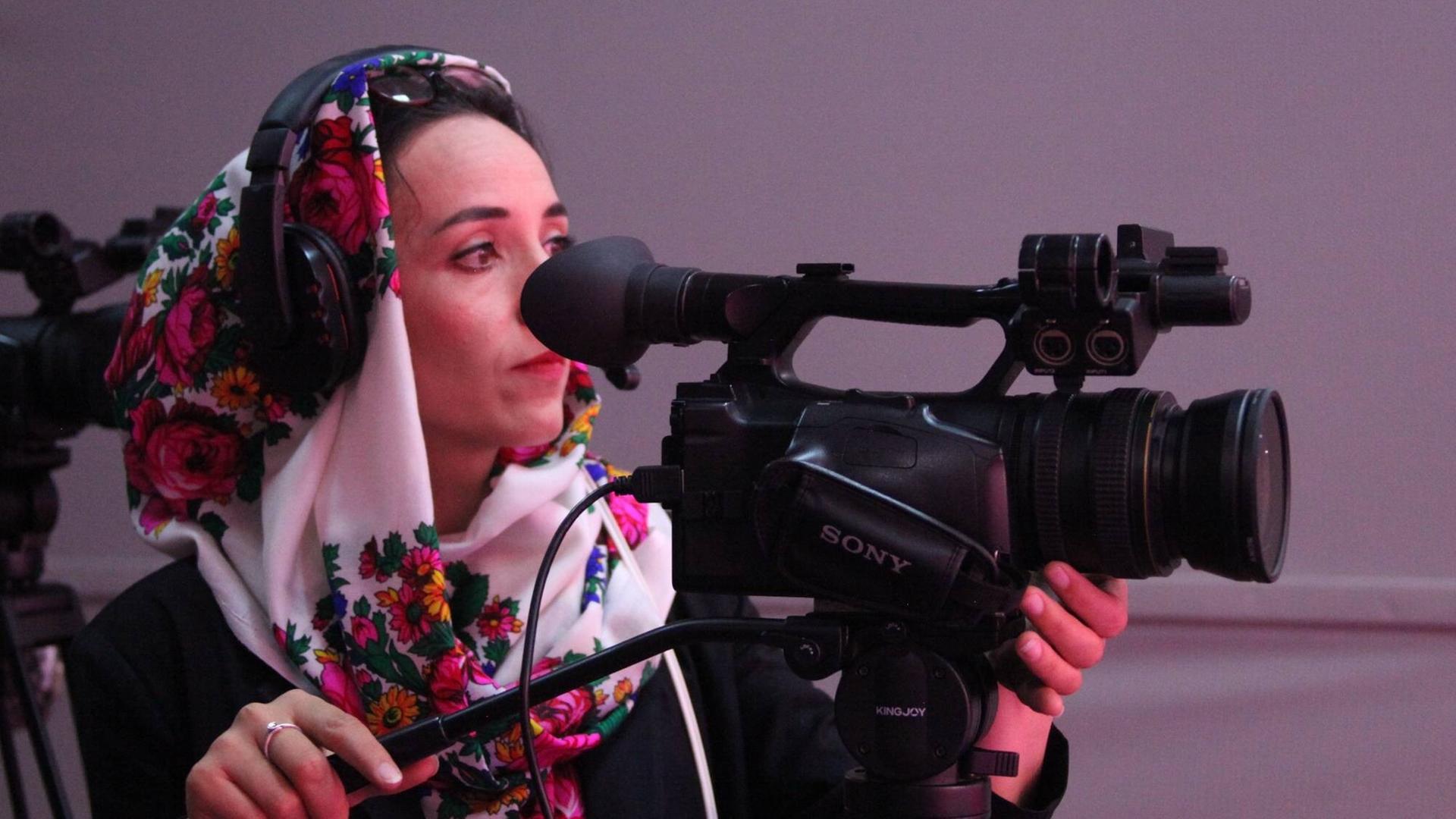 Eine Frau steht an einer TV-Kamera und filmt. Sie trägt ein geblümtes Kopftuch.