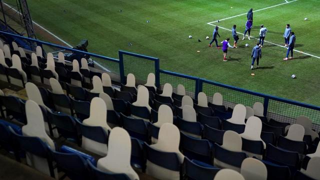 Fußballspieler wärmen sich vor dem Anpfiff auf dem Spielfeld auf. Die dunkle Zuschauertribüne ist leer und mit Pappfiguren anstelle von Zuschauern bestückt.