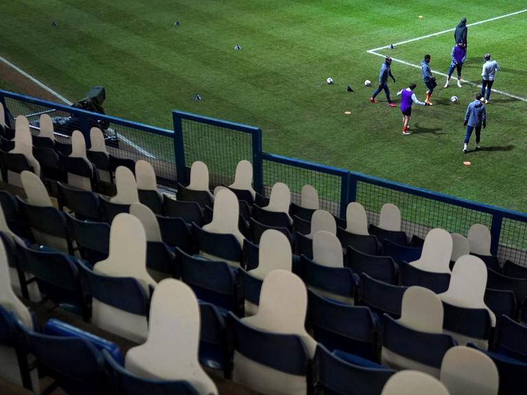 Fußballspieler wärmen sich vor dem Anpfiff auf dem Spielfeld auf. Die dunkle Zuschauertribüne ist leer und mit Pappfiguren anstelle von Zuschauern bestückt.
