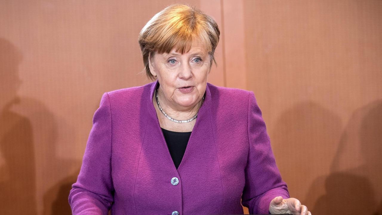 Bundeskanzlerin Angela Merkel steht in einem lila Blazer hinter einer Stuhllehne und spricht zu jemandem, der neben der Kamera steht. 