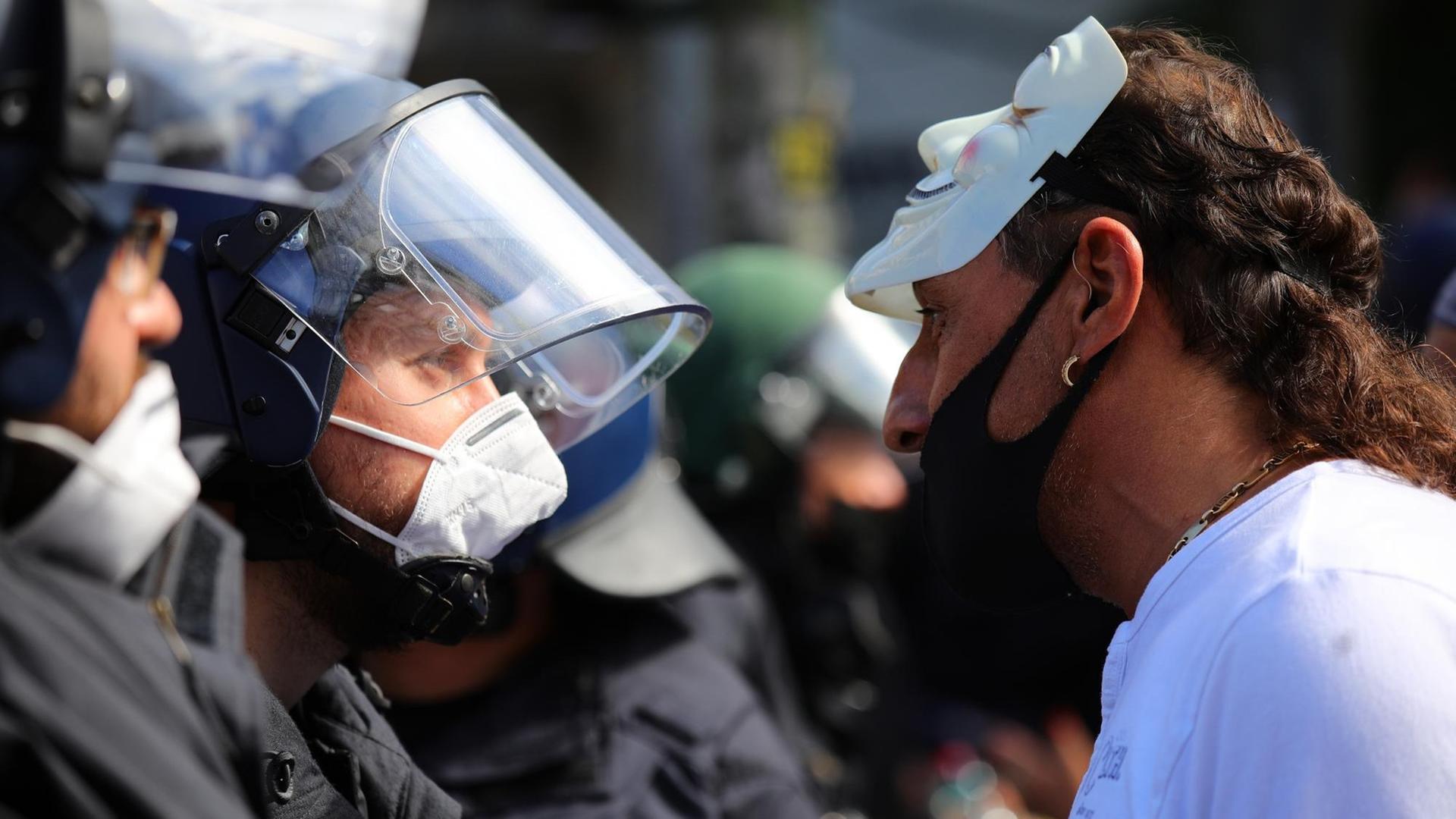 Ein Mann mit Mundschutz unter der Nase und hochgeschobener Anonymous-Maske schreit einen Polizisten an. Ein Mann mit Mundschutz unter der Nase und hochgeschobener Anonymous-Maske schreit einen Polizisten an.