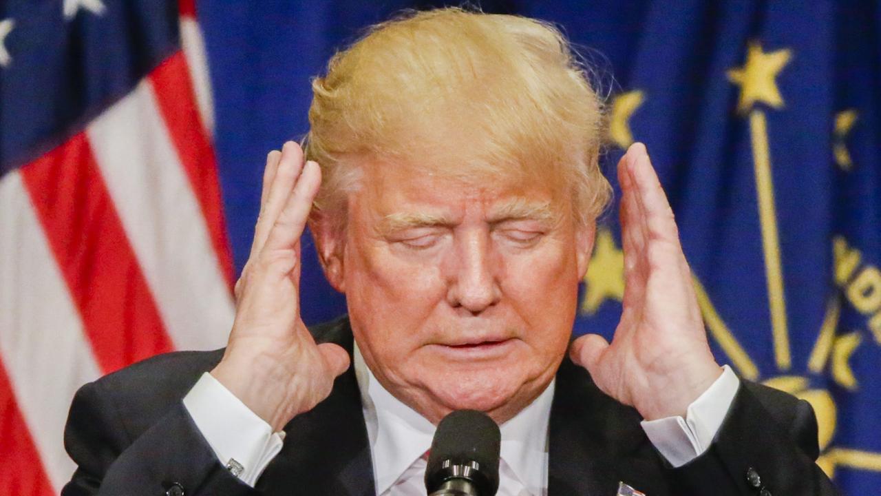 Trump mit geschlossenen Augen vor einer US-Flagge, beide Hände neben den Kopf erhoben.