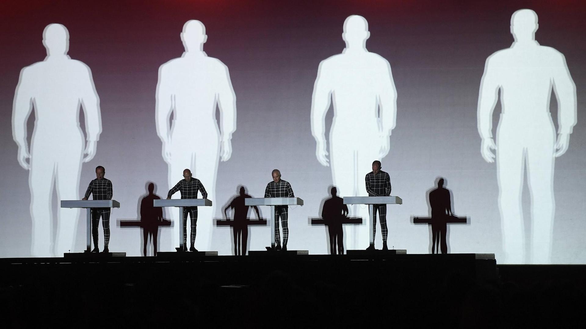 Auf der Bühne stehen die 4 Mitglieder der Band Kraftwerk an Keyboards. Im Hintergrund werden ihre Schatten auf die Leinwand projiziert. Auf der Bühne stehen die 4 Mitglieder der Band Kraftwerk an Keyboards. Im Hintergrund werden ihre Schatten auf die Leinwand projiziert.