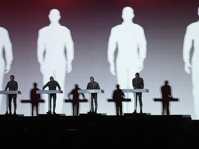 Auf der Bühne stehen die 4 Mitglieder der Band Kraftwerk an Keyboards. Im Hintergrund werden ihre Schatten auf die Leinwand projiziert.