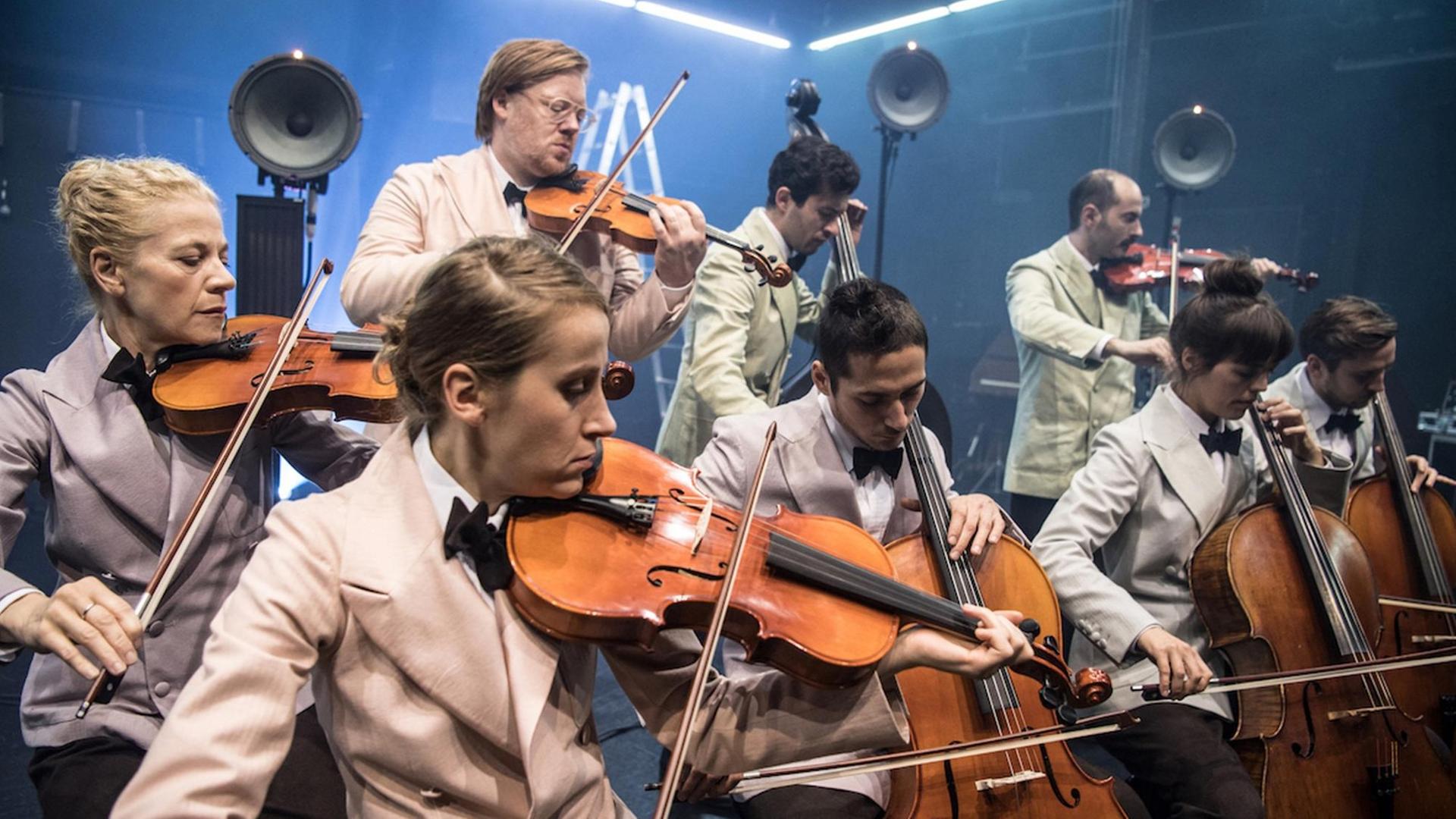 Ein kleines Streichorchester (Geigen und Cello) auf der Bühne. Die Musiker*innen tragen helle Kleidung. Ein kleines Streichorchester (Geigen und Cello) auf der Bühne. Die Musiker*innen tragen helle Kleidung.