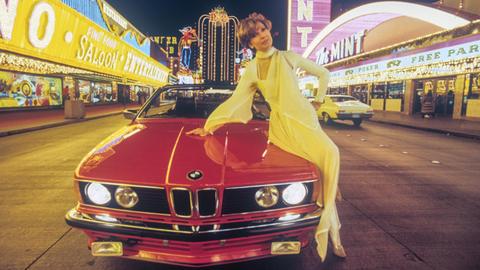 Moderatorin Carolin Reiber 1982 in Las Vegas bei den Dreharbeiten für eine Silvestershow. Sie posiert auf einem roten BMW-Sportwagen, im Hintergrund sind die Lichter der Casinos zu sehen.