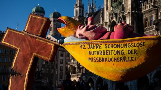 Die Plastik „Der Hängemattenbischof“ steht auf dem Marienplatz. Sie zeigt einen Bischof in einer Hängematte, darauf steht: "12 Jahre schonungslose Aufarbeitung der Missbrauchsfälle!" und ist an einem gebrochenen Kreiz aufgehängt

