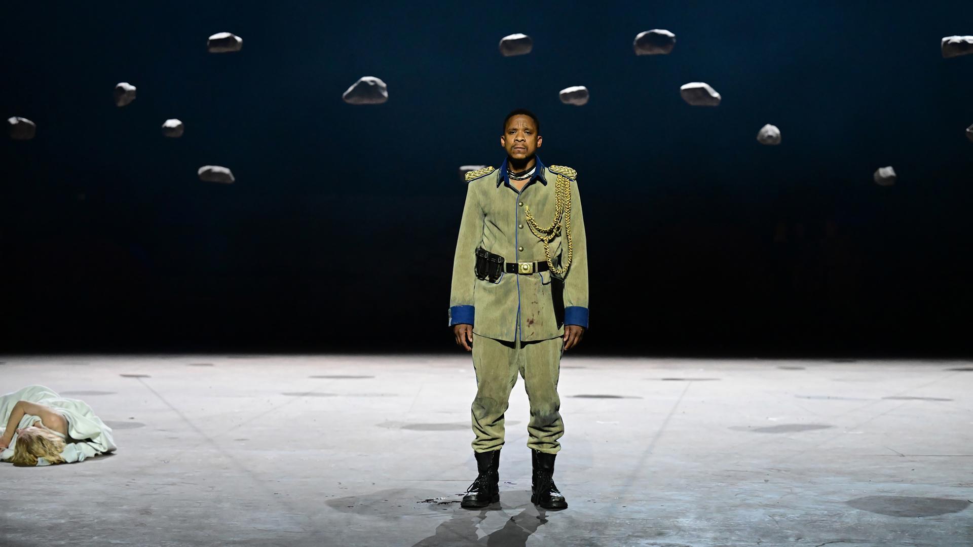 Schauspieler Bongile Mantsai steht in einer zerschlissenen altmodischen Militäruniform auf der Bühne. Am linken Bildrand liegt eine blonde Frau auf dem Boden. Schauspieler Bongile Mantsai steht in einer zerschlissenen altmodischen Militäruniform auf der Bühne. Am linken Bildrand liegt eine blonde Frau auf dem Boden.