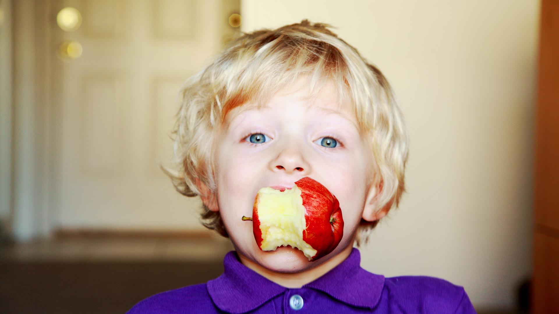 Ein Kind hält einen angebissenen Apfel im Mund (ohne Hände).