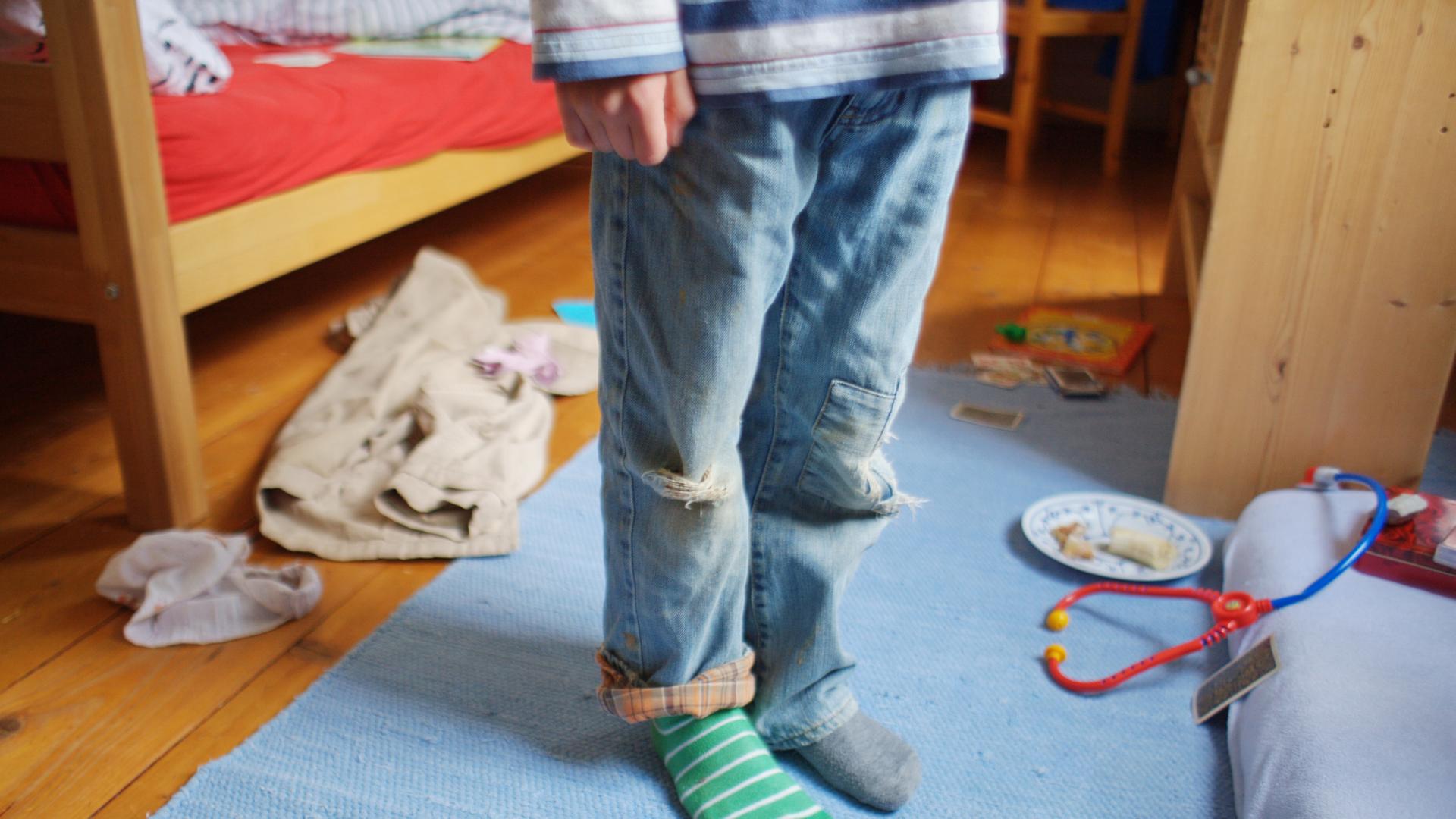 Ein Kind steht mit schmutziger Kleidung, zerrissenen Hosen und verschiedenen Strümpfen in einem unaufgräumten Zimmer. Ein Kind steht mit schmutziger Kleidung, zerrissenen Hosen und verschiedenen Strümpfen in einem unaufgräumten Zimmer.