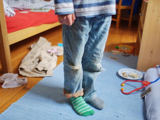 Ein Kind steht mit schmutziger Kleidung, zerrissenen Hosen und verschiedenen Strümpfen in einem unaufgräumten Zimmer, in dem Kleidungsstücke, billiges Spielzeug und Essensreste auf dem Boden liegen (gestellte Szene). 