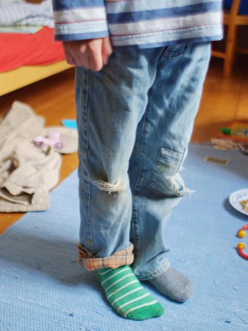 Ein Kind steht mit schmutziger Kleidung, zerrissenen Hosen und verschiedenen Strümpfen in einem unaufgräumten Zimmer, in dem Kleidungsstücke, billiges Spielzeug und Essensreste auf dem Boden liegen (gestellte Szene). 