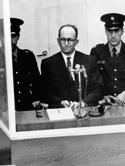 Der NS-Kriegsverbrecher Adolf Eichmann während seiner Vernehmung am ersten Prozesstag vor dem Jerusalemer Bezirksgericht am 11.4.1961. Vor ihm steht ein Mikrofon, neben ihm zwei Wachen.