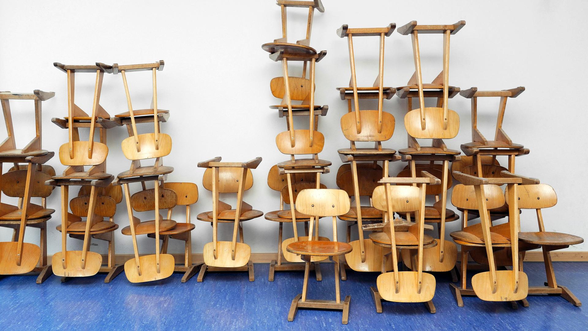 Stapel alter Holzstühle in einer Schule, die auf einem blauen Linoleumboden an einer Wand stehen. Stapel alter Holzstühle in einer Schule, die auf einem blauen Linoleumboden an einer Wand stehen.