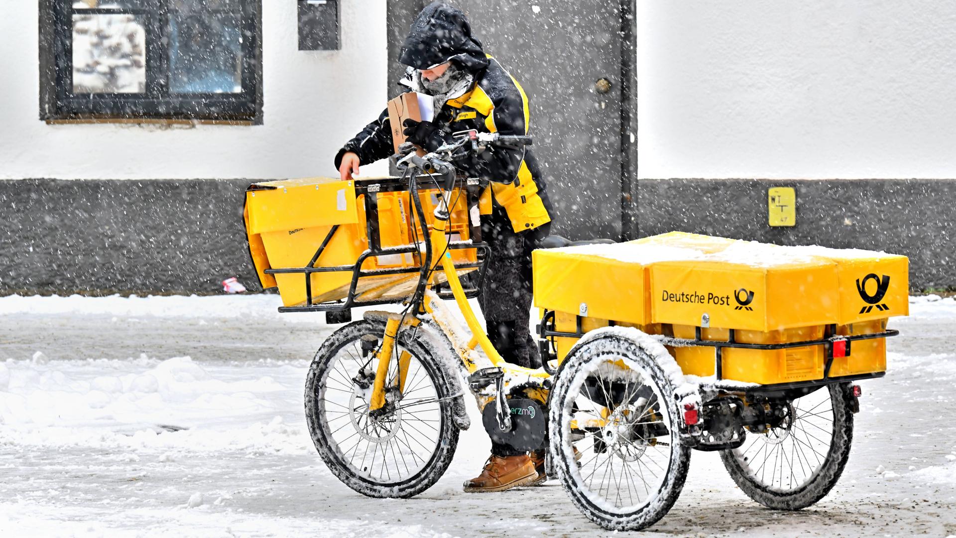 Eine Postzustellerin mit ihrem gelben Lastenfahrrad an einem schneereichen Wintertag in Bad Reichenhall bei der Arbeit. Sie trägt im Schneetreiben die Post aus. Eine Postzustellerin mit ihrem gelben Lastenfahrrad an einem schneereichen Wintertag in Bad Reichenhall bei der Arbeit. Sie trägt im Schneetreiben die Post aus.