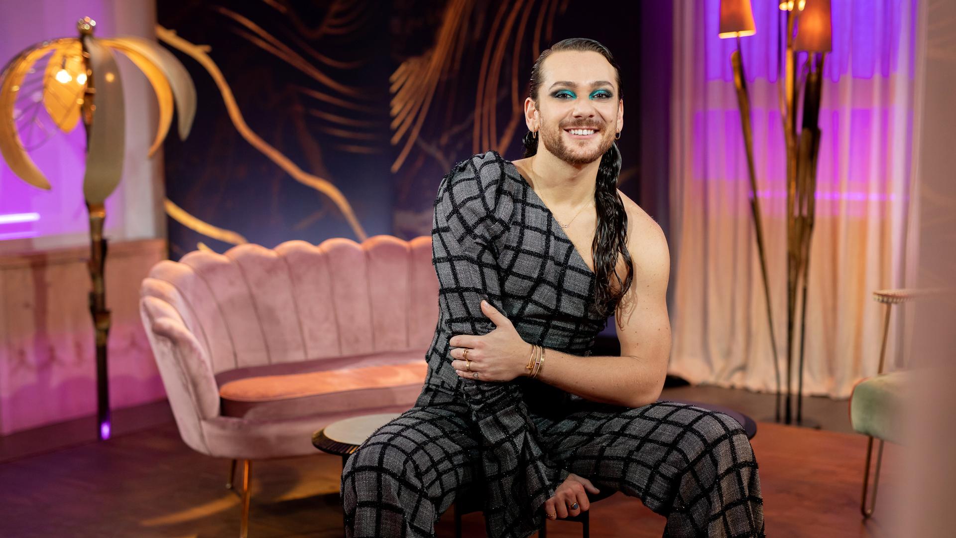 Blick auf Riccardo Simonetti in den Kulissen seiner Show. Er sitz im Vordergrund auf einem kleinen Tisch, im Hintergrund ein rosa Sofa, kleine Lampen und Gardinen vor den Fenstern. Blick auf Riccardo Simonetti in den Kulissen seiner Show. Er sitz im Vordergrund auf einem kleinen Tisch, im Hintergrund ein rosa Sofa, kleine Lampen und Gardinen vor den Fenstern.