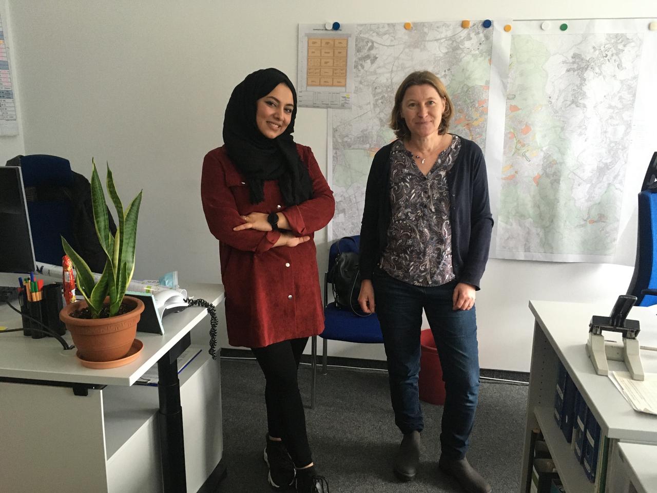 Fathemah steht neben ihrer Chefin im Büro. Sie trägt Turnschuh und Kopftuch. Beide lächeln in die Kamera.