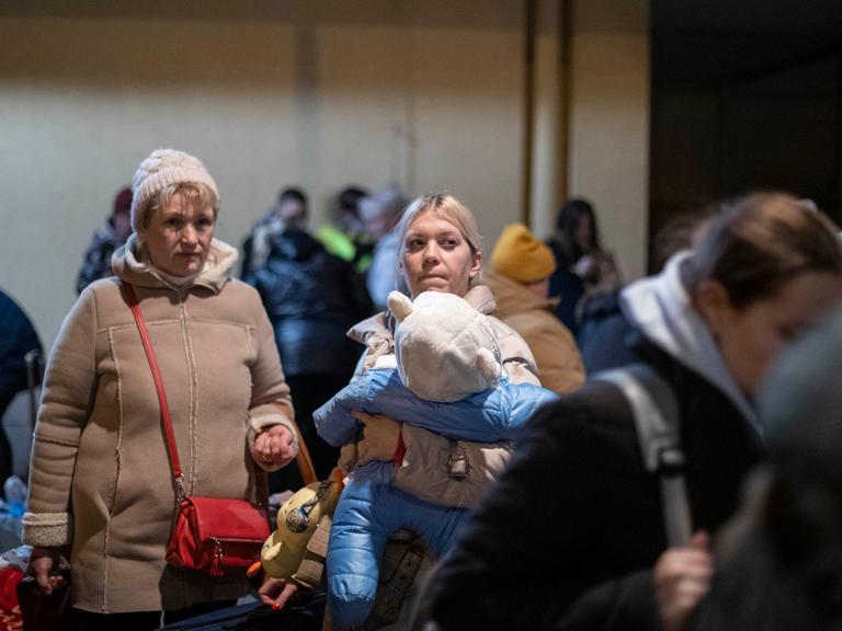 Zwei weiße, blonde Frauen in Winterkleidung stehen zwischen Gepäck und anderen Frauen an vermutlich einem Bahnhof. Die rechte der beiden Frauen hat ein kleines Kind auf dem Arm.