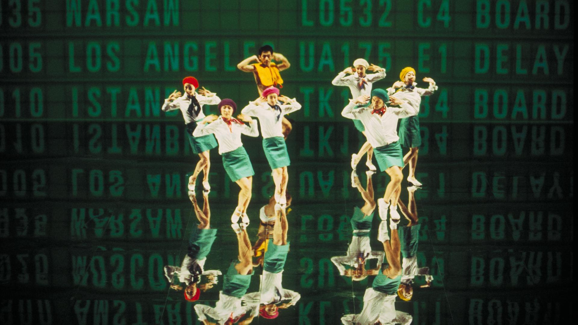 Sieben Personen, die an Flugbegleiter erinnen, stehen vor einer Leinwand. An der Leinwand sind in großen Buchstaben Flugnummern und Ziele angeschlagen. Das Bild ist Teil einer Videoinstallation eines japanischen Künstlerkollektivs.