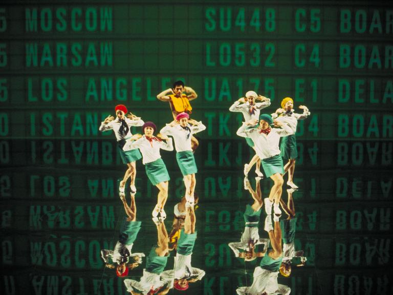 Sieben Personen, die an Flugbegleiter erinnen, stehen vor einer Leinwand. An der Leinwand sind in großen Buchstaben Flugnummern und Ziele angeschlagen. Das Bild ist Teil einer Videoinstallation eines japanischen Künstlerkollektivs.