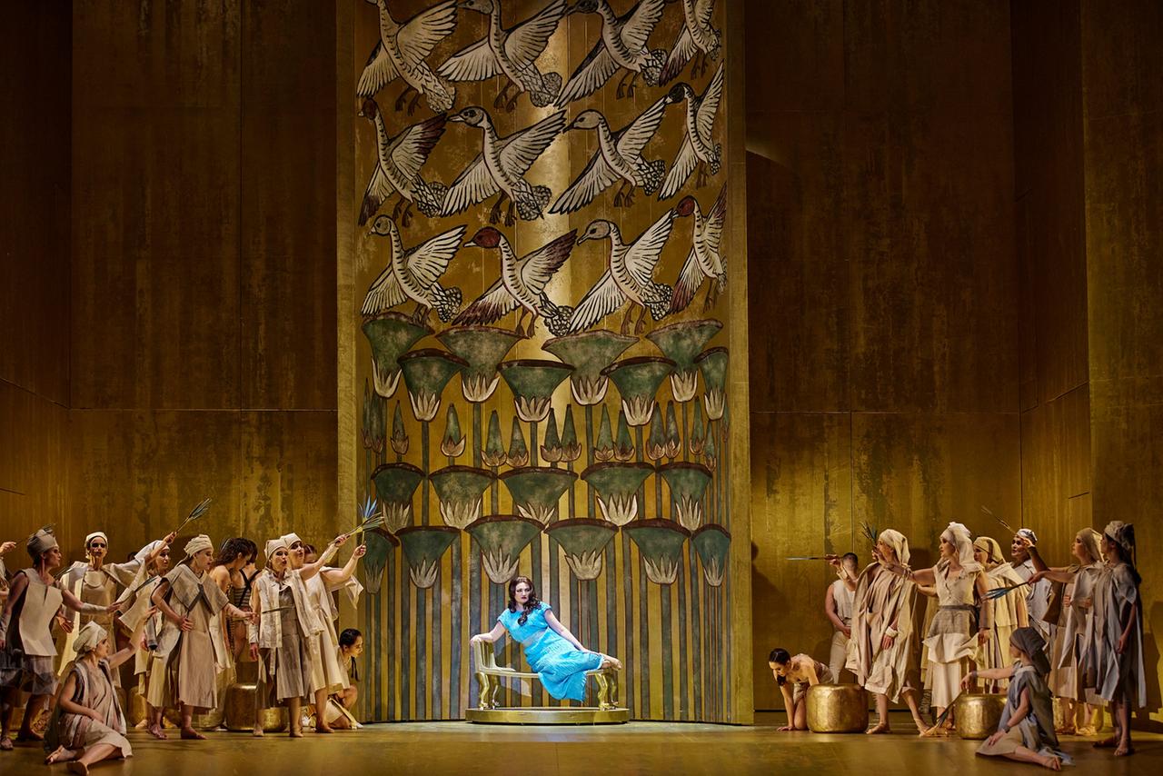 Eine Frau in einem türkis blauen Kleid liegt in einer herrschaftlichen Position auf einem goldenen Thron. Hinter ihr verzierte Wände, neben ihr als alte Ägypter erkennbare Menschen.