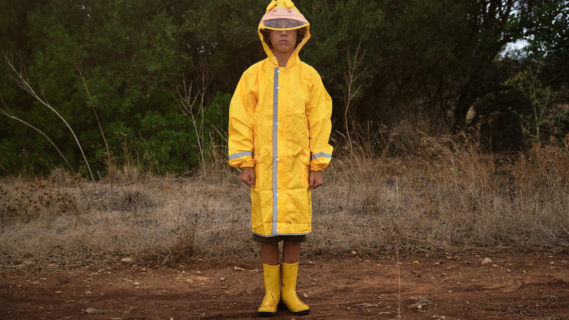 Ein Junge mit gelber Regenkleidung steht allein in einer trockenen Parklandschaft Ein Junge mit gelber Regenkleidung steht allein in einer trockenen Parklandschaft