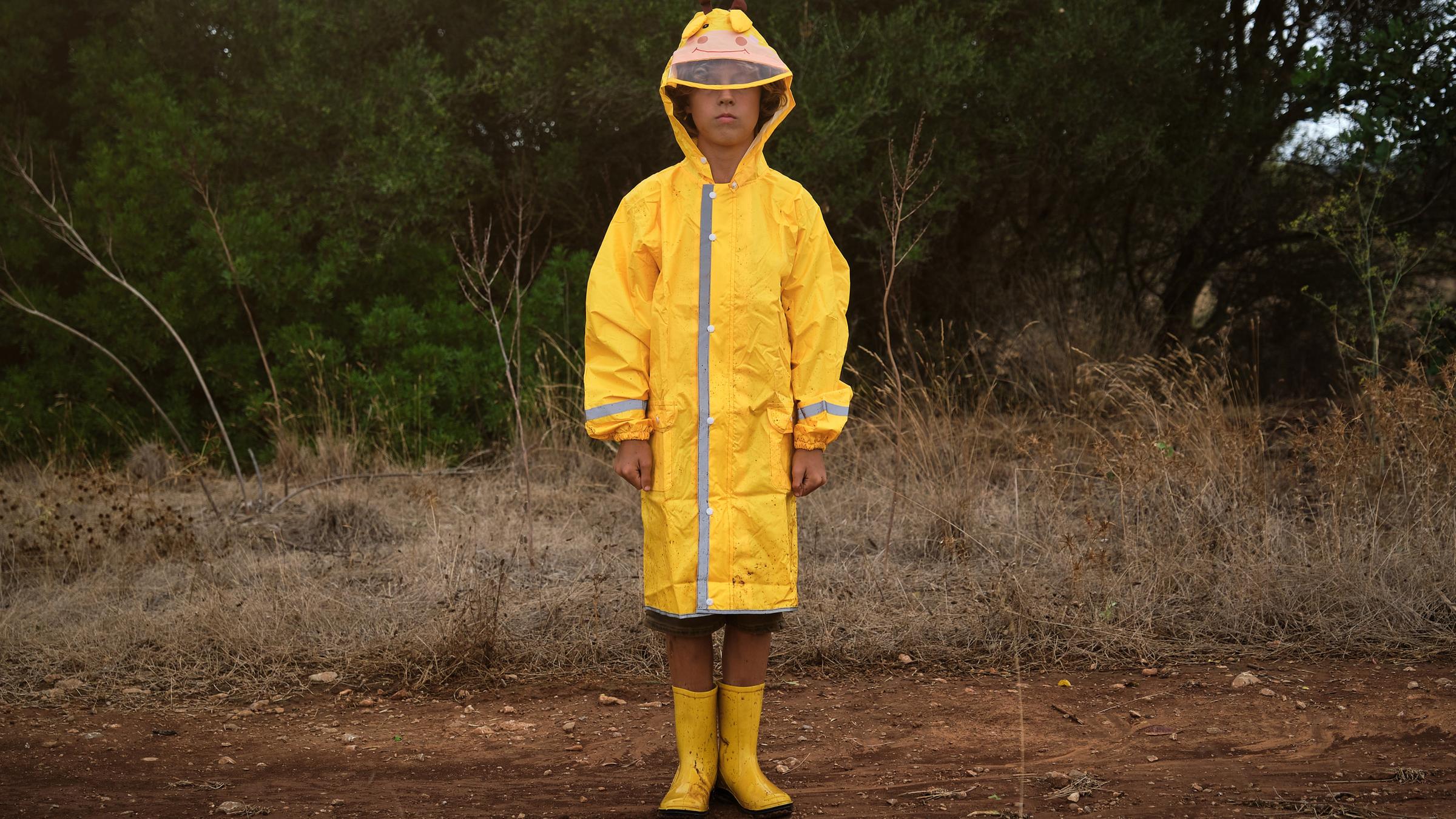 Ein Junge mit gelber Regenkleidung steht allein in einer trockenen Parklandschaft