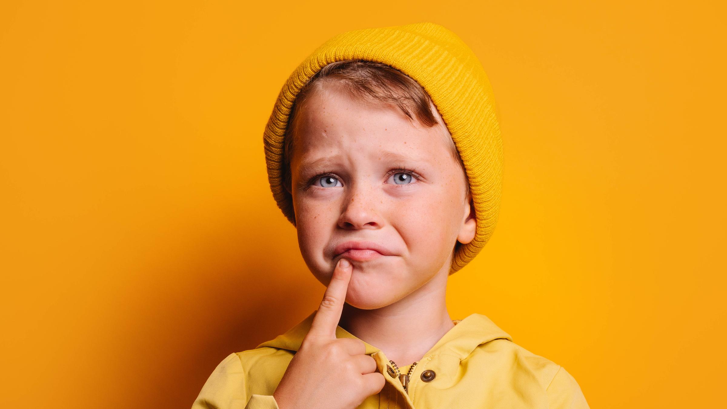 Ein Kind mit gelber Mütze vor gelbem Hintergrund mit grübelndem Gesichtsausdruck und dem Zeigefinger am Kinn Ein Kind mit gelber Mütze vor gelbem Hintergrund mit grübelndem Gesichtsausdruck und dem Zeigefinger am Kinn