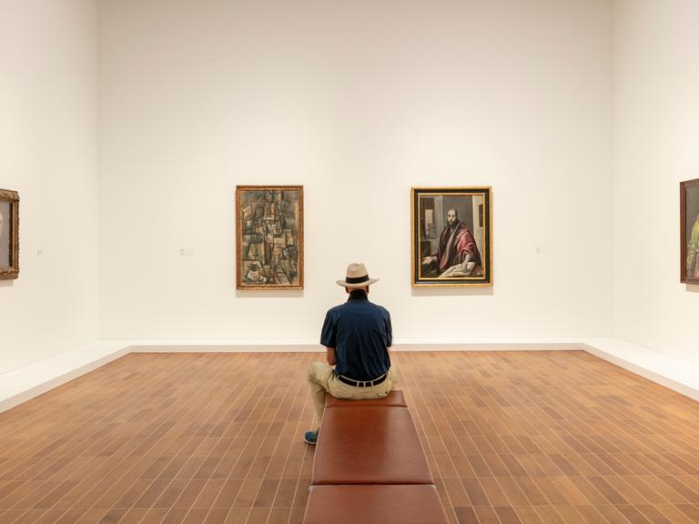 Ausstellungsansicht: Ein Mann sitzt auf einer Bank und betrachtet die Werke von Picasso und El Greco.