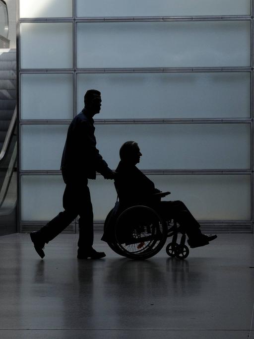 Ein junger Mann schiebt einen Rollstuhlfahrer durch eine Halle. Es könnte eine Flughafenhalle sein. Von beiden ist nur die Silhouette erkennbar. 