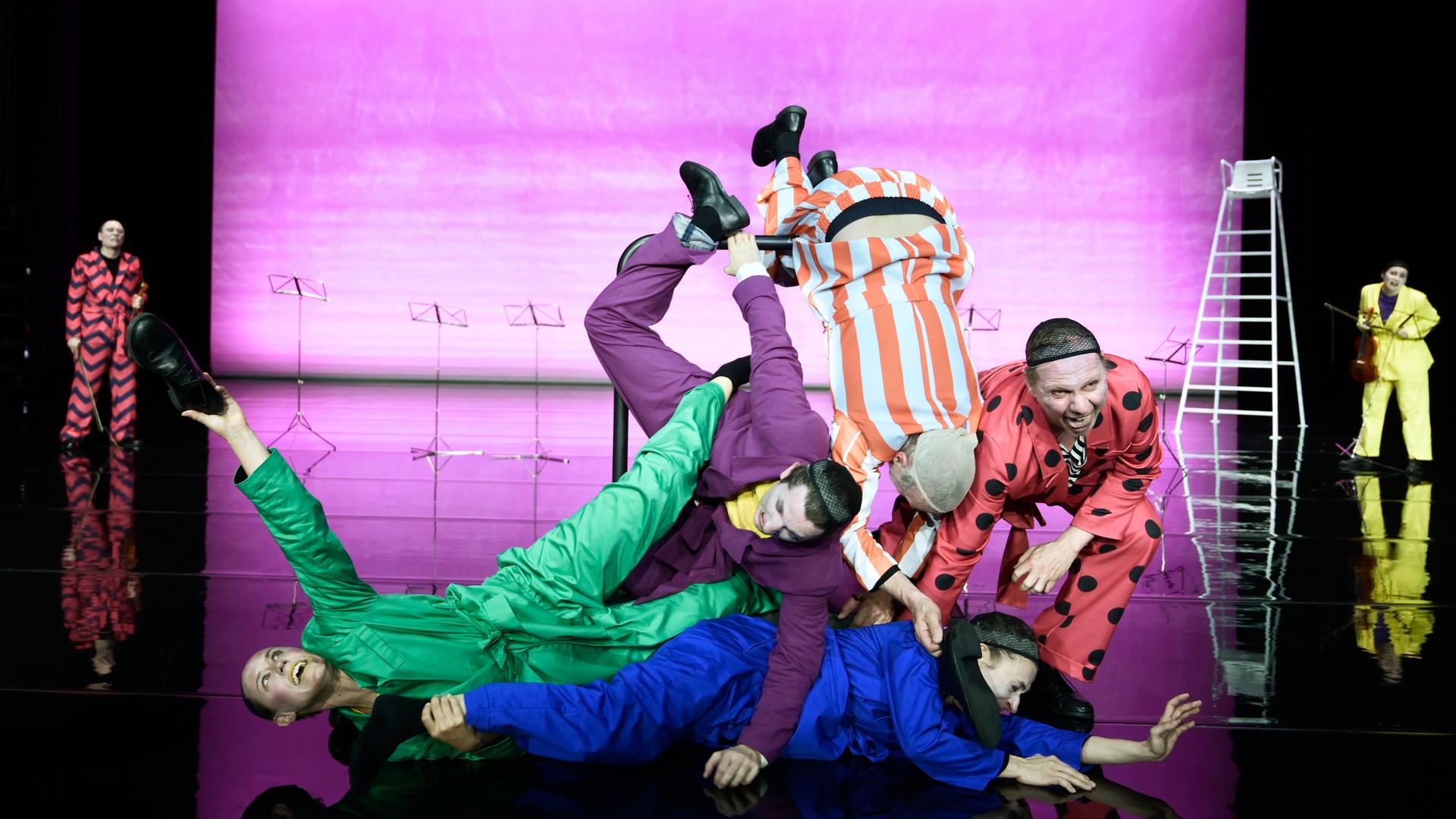 Fünf Clowns in bunten Overalls formen in der Bühnenmitte eine menschliche Pyramide. Dahinter steht links ein Mann mit Geige, rechts eine Frau mit Geige. Im Hintergrund stehen Notenständer und ein Bademeisterstuhl. Fünf Clowns in bunten Overalls formen in der Bühnenmitte eine menschliche Pyramide. Dahinter steht links ein Mann mit Geige, rechts eine Frau mit Geige. Im Hintergrund stehen Notenständer und ein Bademeisterstuhl.