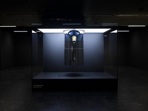 Ein futuristisch anmutendes Gebilde in einem Kasten steht in einem schwarzen Raum. Darauf steht der Name des Quantencomputer "IBM Quantum System One".