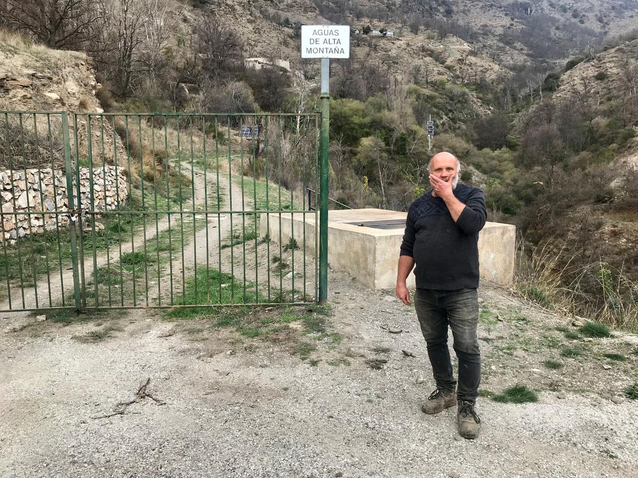Ein älterer Mann mit Glatze und weißem Bart steht in einer bergigen Landschaft vor einem Gittertor. Ein älterer Mann mit Glatze und weißem Bart steht in einer bergigen Landschaft vor einem Gittertor.