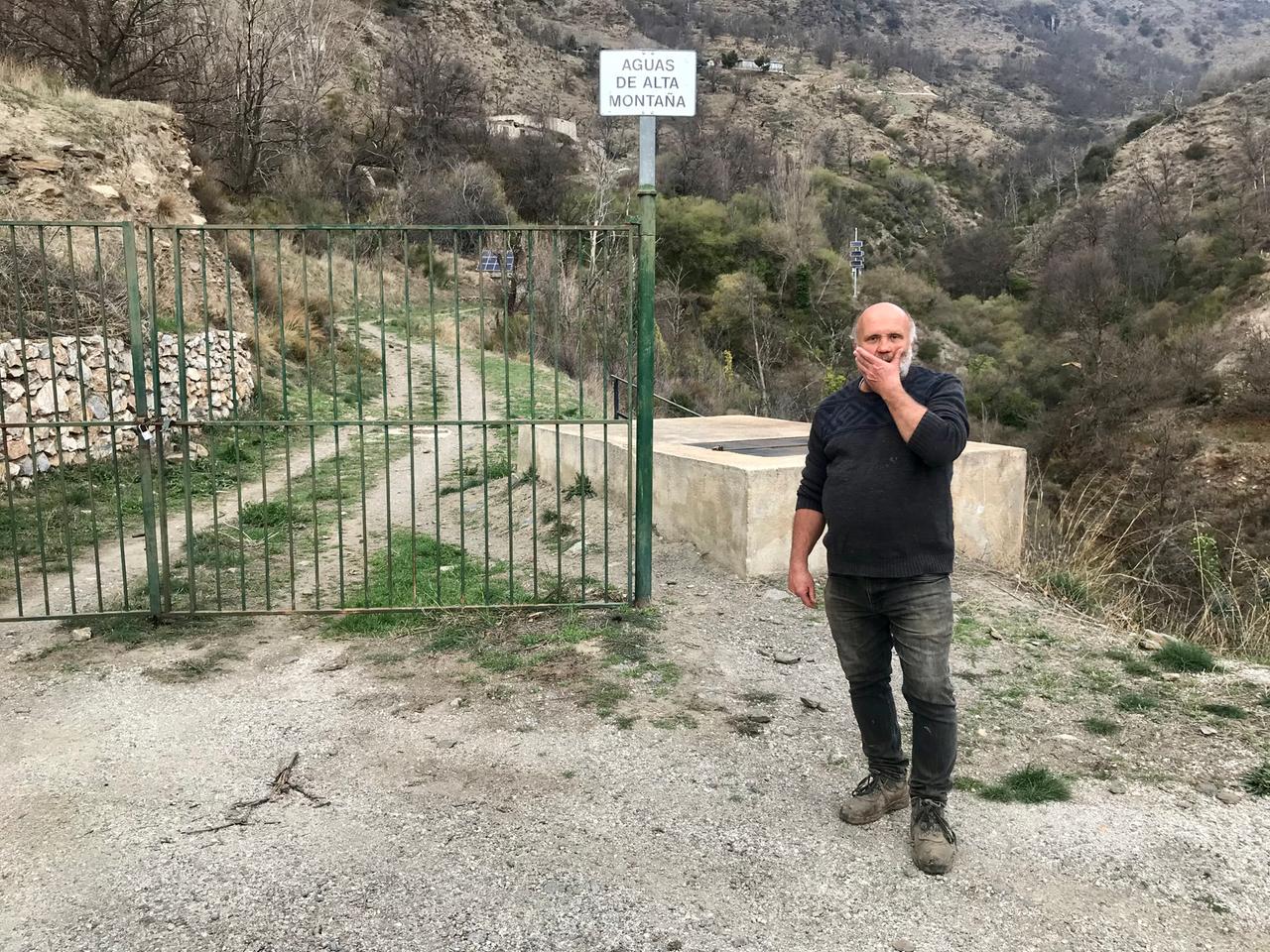 Ein älterer Mann mit Glatze und weißem Bart steht in einer bergigen Landschaft vor einem Gittertor.