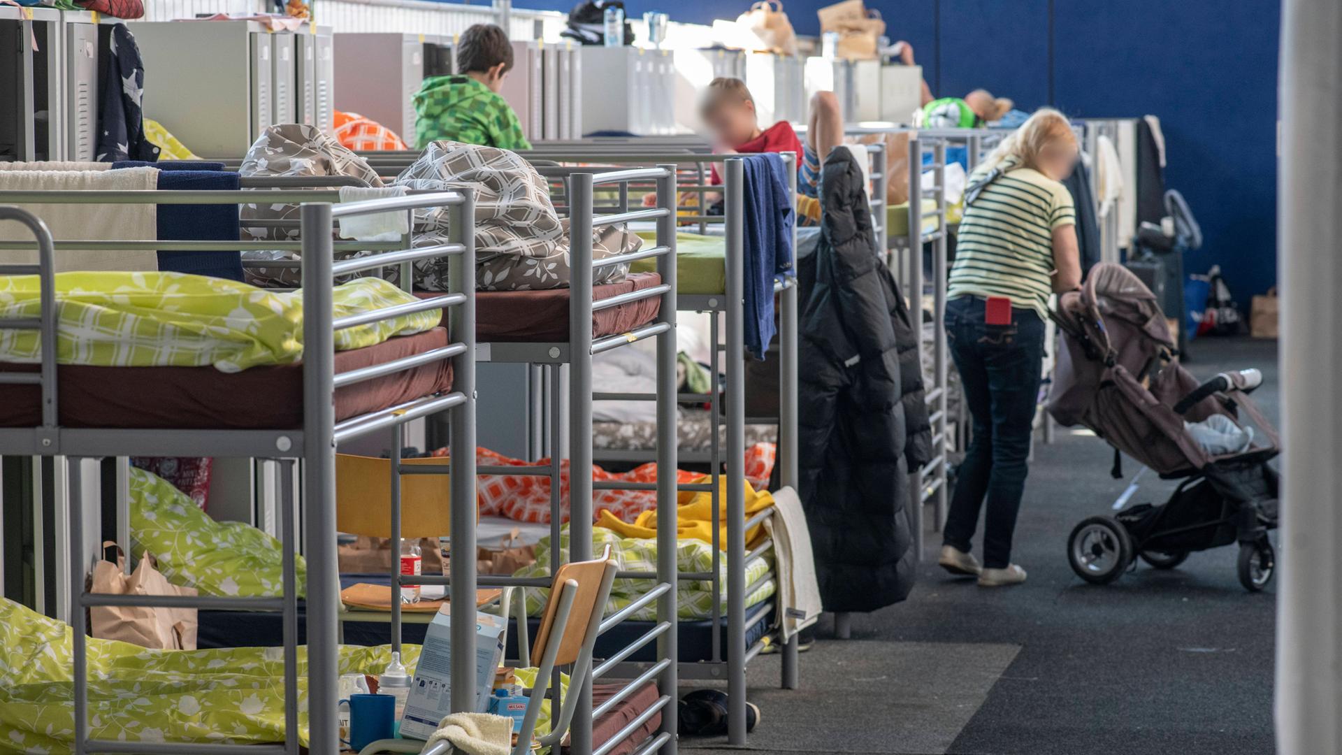 In einer Flüchtlingsunterkunft stehen Doppelstockbetten, auf denen zwei Kinder sitzen, daneben schiebt eine junge Frau einen Kinderwagen. In einer Flüchtlingsunterkunft stehen Doppelstockbetten, auf denen zwei Kinder sitzen, daneben schiebt eine junge Frau einen Kinderwagen.