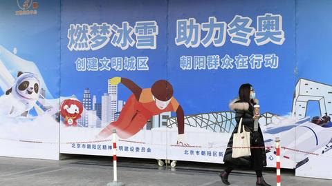 Eine Frau im Wintermantel und mit Einkaufstüten läuft an einer großen Werbetafel für die Olympischen Winterspiele in Peking vorbei.