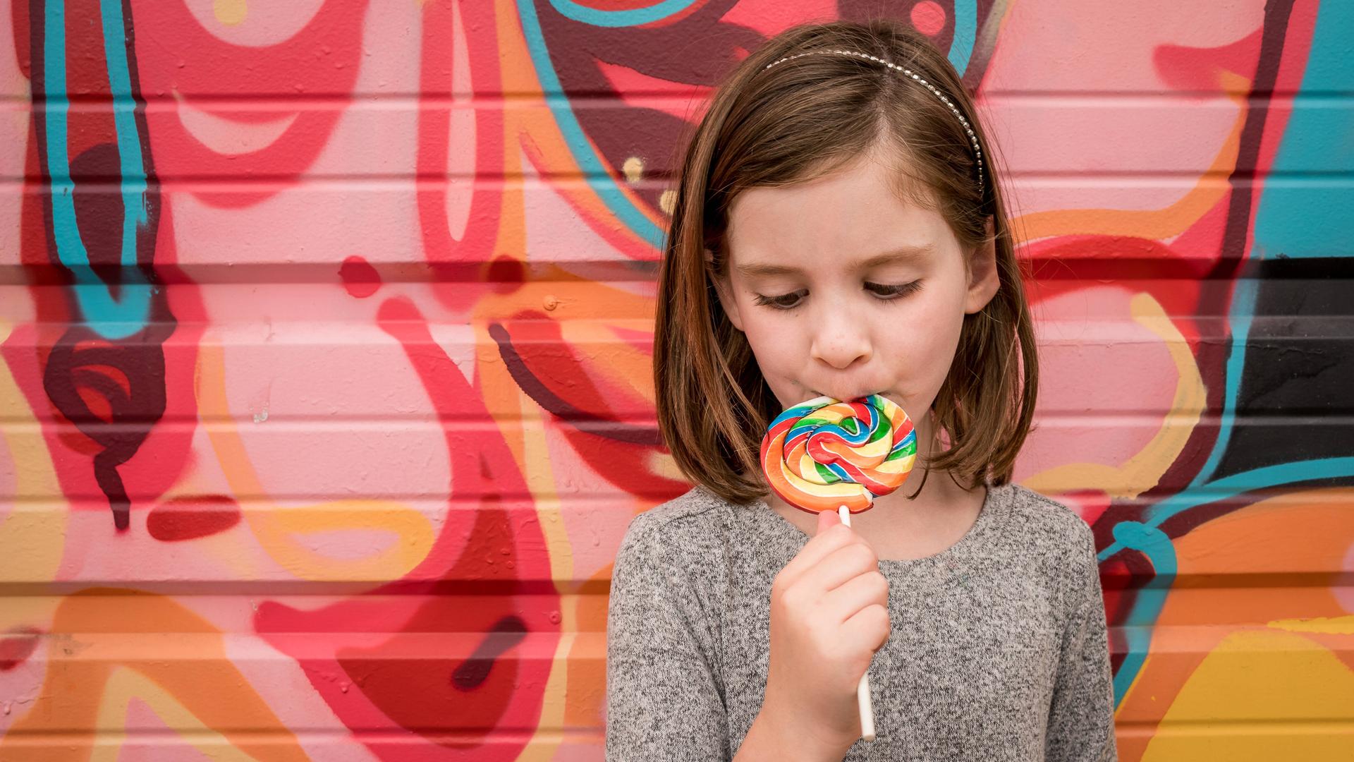 Ein Mädchen steht mit einem farbenfrohen Lolli vor einer bunten Wand Ein Mädchen steht mit einem farbenfrohen Lolli vor einer bunten Wand