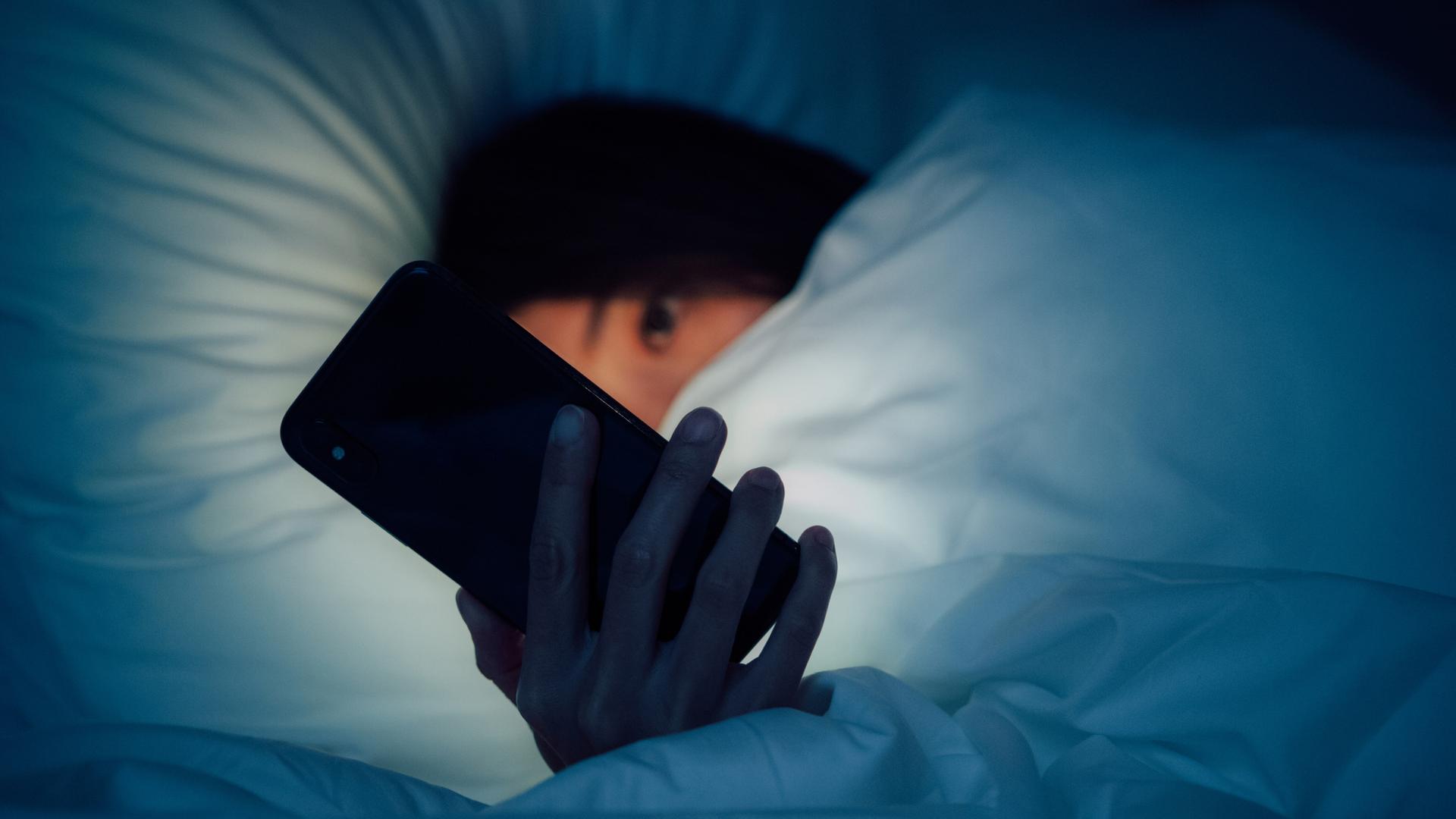 Zu sehen ist eine Frau, eingehüllt in eine dicke Bettdecke. Sie blickt auf ein leuchtendes Smartphone, dass ihr besorgtes Gesicht im Dunkeln erhellt. Zu sehen ist eine Frau, eingehüllt in eine dicke Bettdecke. Sie blickt auf ein leuchtendes Smartphone, dass ihr besorgtes Gesicht im Dunkeln erhellt.