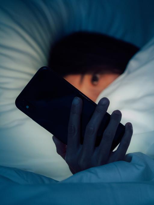 Zu sehen ist eine Frau, eingehüllt in eine dicke Bettdecke. Sie blickt auf ein leuchtendes Smartphone, dass ihr besorgtes Gesicht im Dunkeln erhellt.