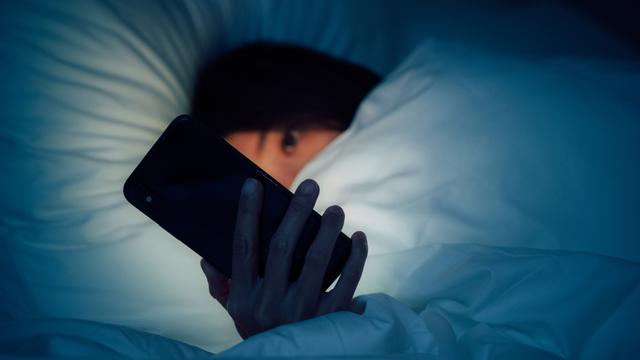 Zu sehen ist eine Frau, eingehüllt in eine dicke Bettdecke. Sie blickt auf ein leuchtendes Smartphone, dass ihr besorgtes Gesicht im Dunkeln erhellt.