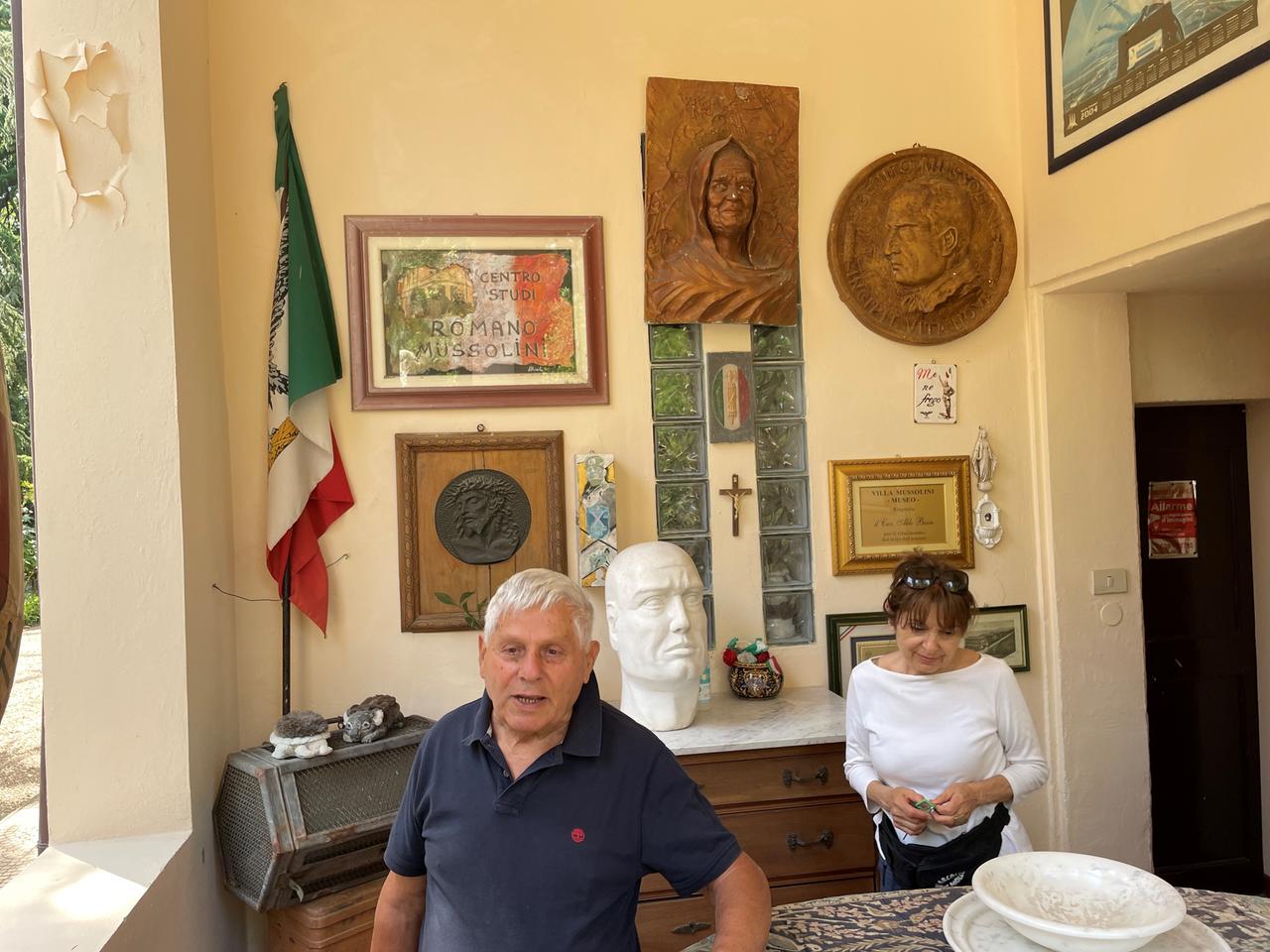 Ein älteres Paar steht vor einem Tisch in einem Raum, der mit Mussolini-Erinnerungsstücken, Bildern, Büsten gefüllt ist.