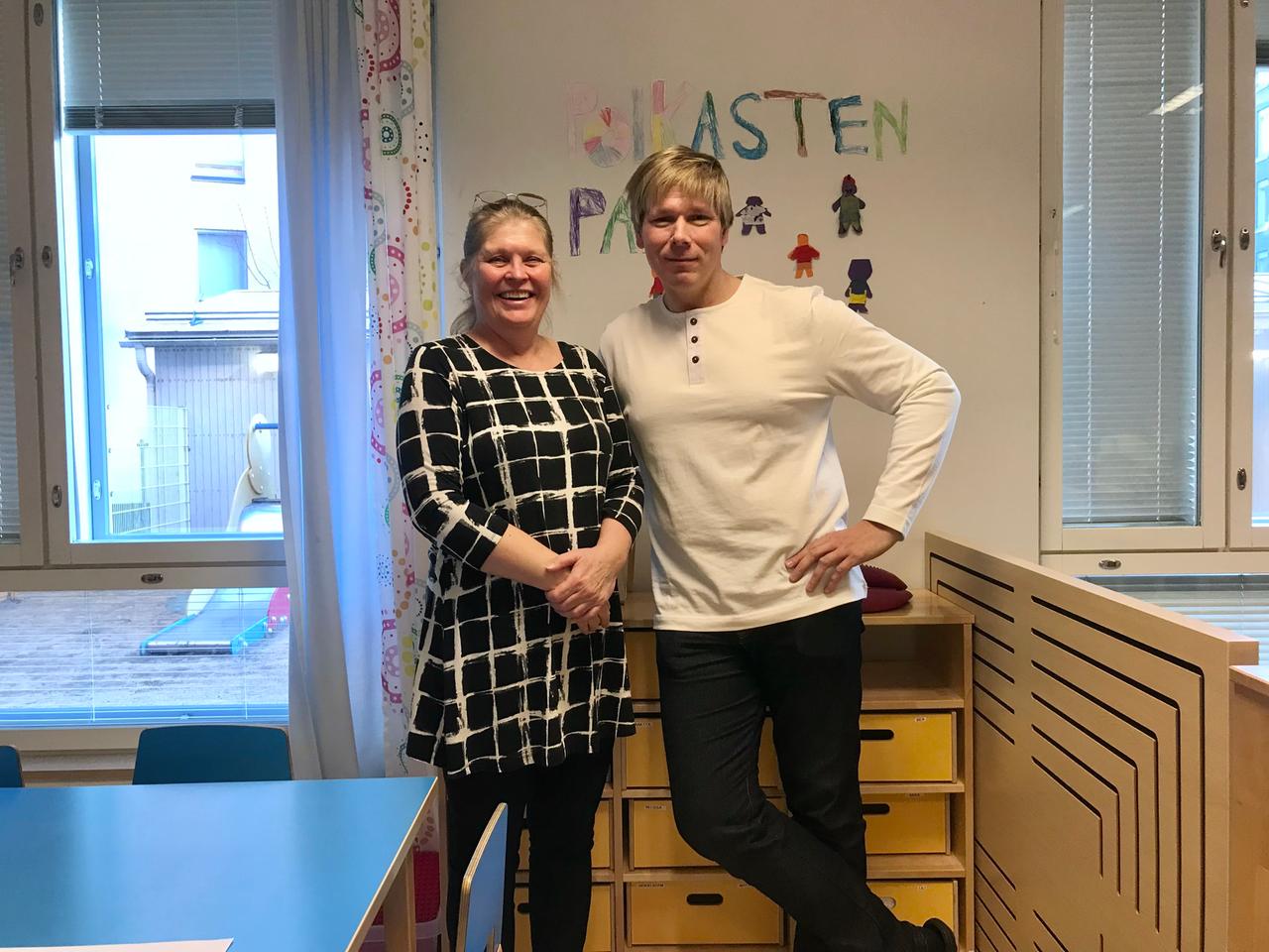 Eine ältere Frau im schwarz-weißen Kleidchen und ein junger Mann im weißen Shirt stehen nebeneinander vor einer bunten Kindergartenwand in einem Raum mit Einrichtung aus Holz. Eine ältere Frau im schwarz-weißen Kleidchen und ein junger Mann im weißen Shirt stehen nebeneinander vor einer bunten Kindergartenwand in einem Raum mit Einrichtung aus Holz.