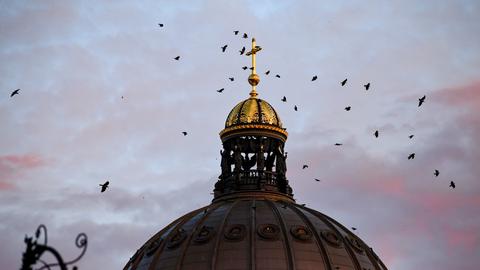 Krähen überfliegen am Abend das Kreuz der Kuppel am Humboldt Forum. 