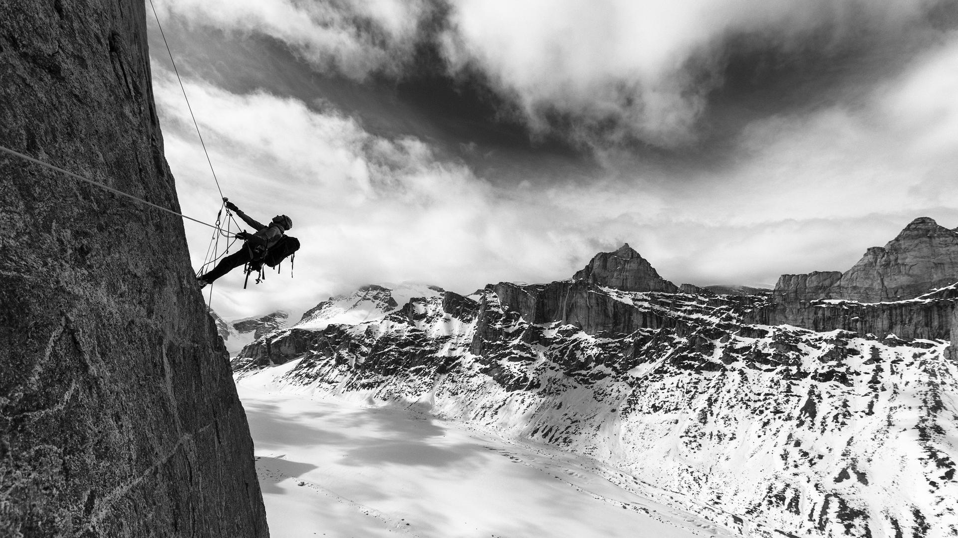 Eine Schwarz-Weiß-Aufnahme zeigt einen Bergsteiger, angeseilt an einer steilen Felswand, Baffin Island (2016). Eine Schwarz-Weiß-Aufnahme zeigt einen Bergsteiger, angeseilt an einer steilen Felswand, Baffin Island (2016).