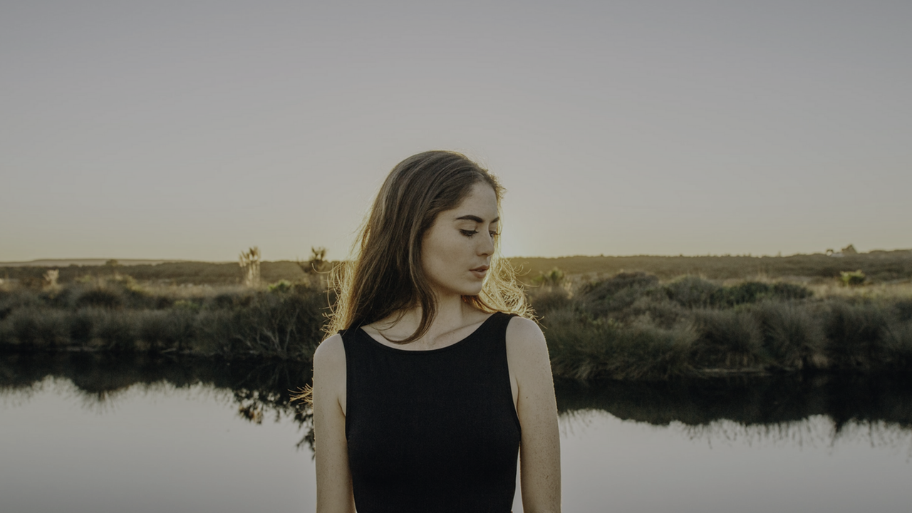 Eine junge Frau, den Kopf zur Seite geneigt, steht vor einem kleinen See. Im Hintergrund ist eine Wiesenlandschaft zu sehen.