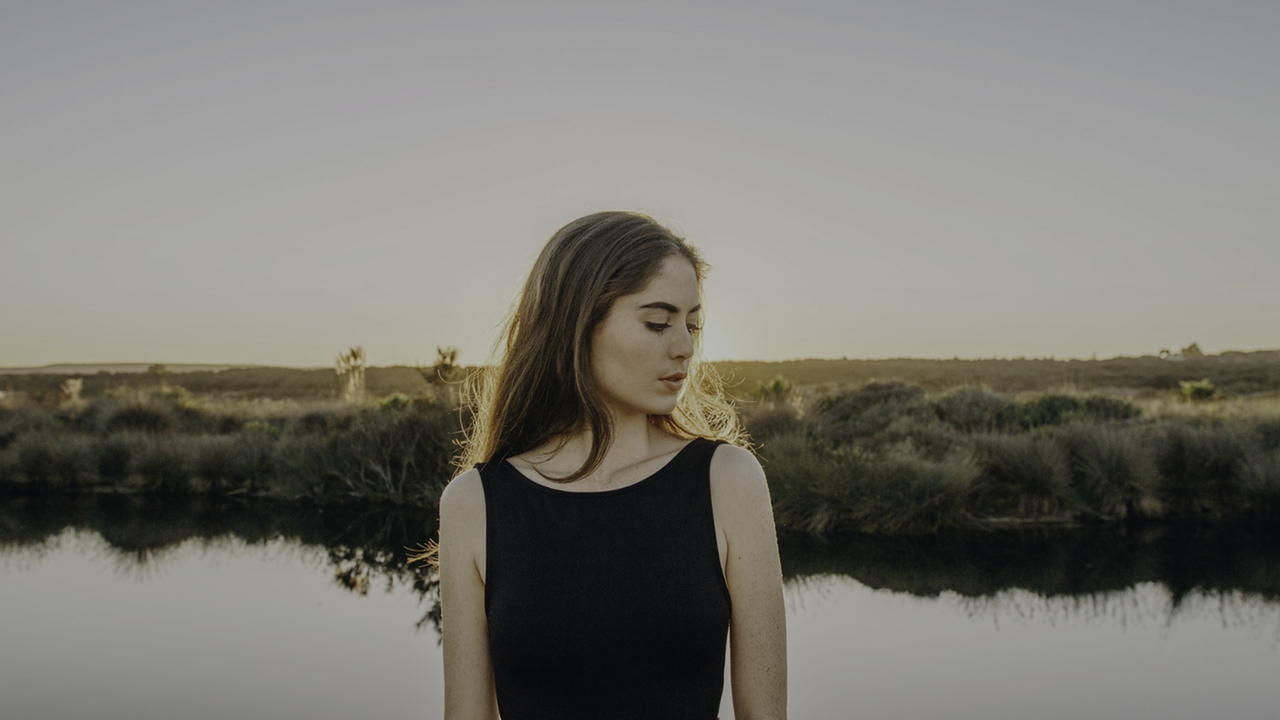 Eine junge Frau, den Kopf zur Seite geneigt, steht vor einem kleinen See. Im Hintergrund ist eine Wiesenlandschaft zu sehen. Eine junge Frau, den Kopf zur Seite geneigt, steht vor einem kleinen See. Im Hintergrund ist eine Wiesenlandschaft zu sehen.