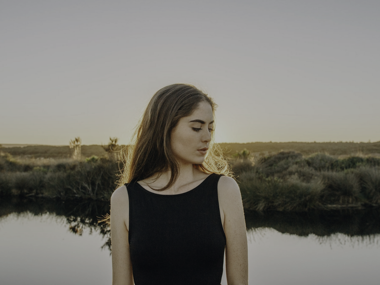 Eine junge Frau, den Kopf zur Seite geneigt, steht vor einem kleinen See. Im Hintergrund ist eine Wiesenlandschaft zu sehen.